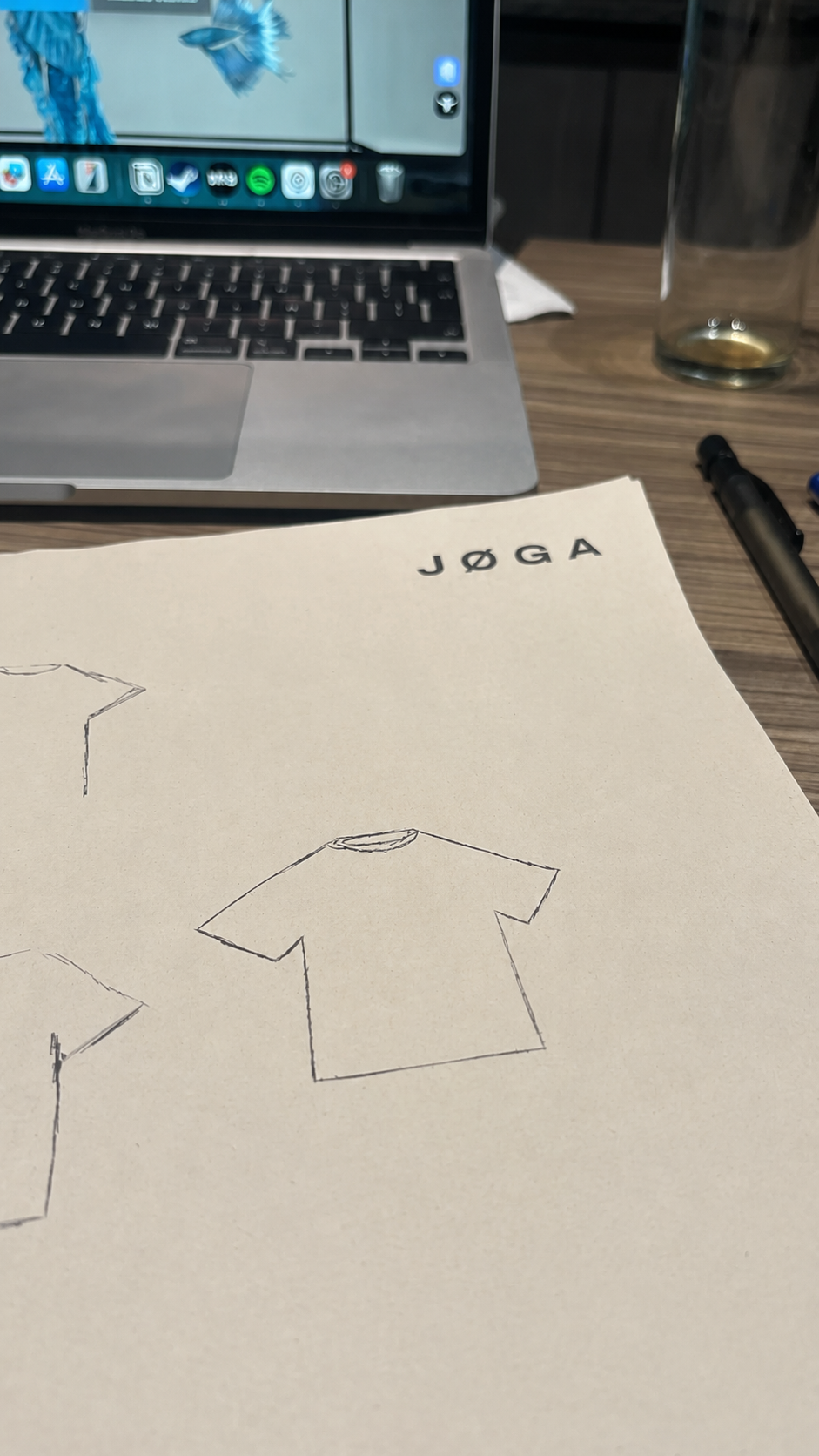 Early JØGA tee sketches on paper next to a laptop